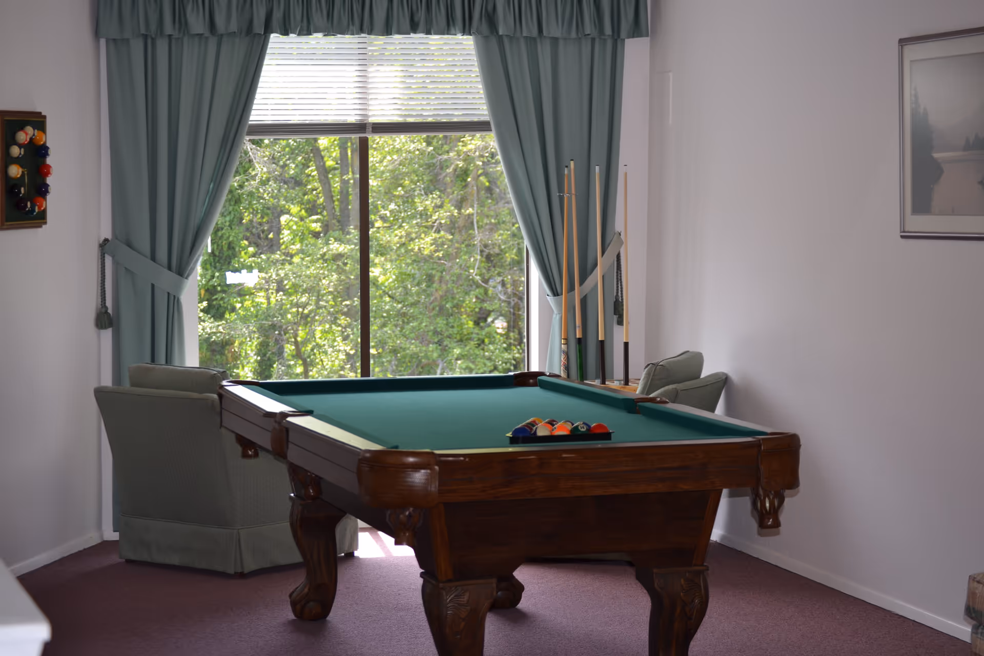 A room with a pool table set up for a game, two green armchairs, a large window with green curtains, and a rack holding pool cues. Outside the window, green trees are visible.