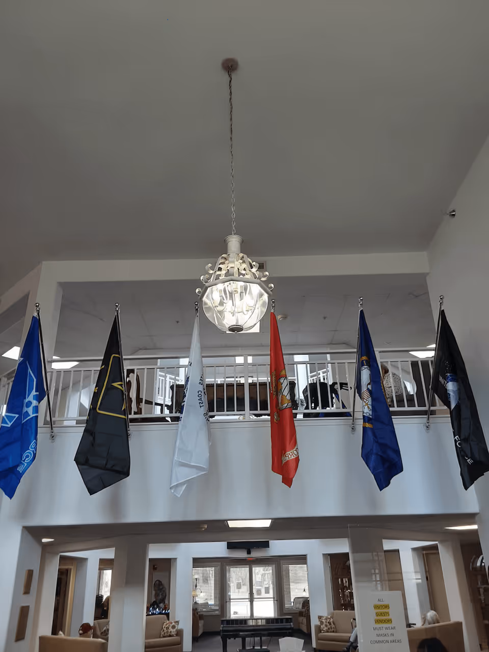 Interior view of a senior living facility lobby area with six flags hanging from the upper balcony railing. A decorative chandelier hangs from the ceiling. Below, there is a seating area with couches and a piano near the entrance doors.