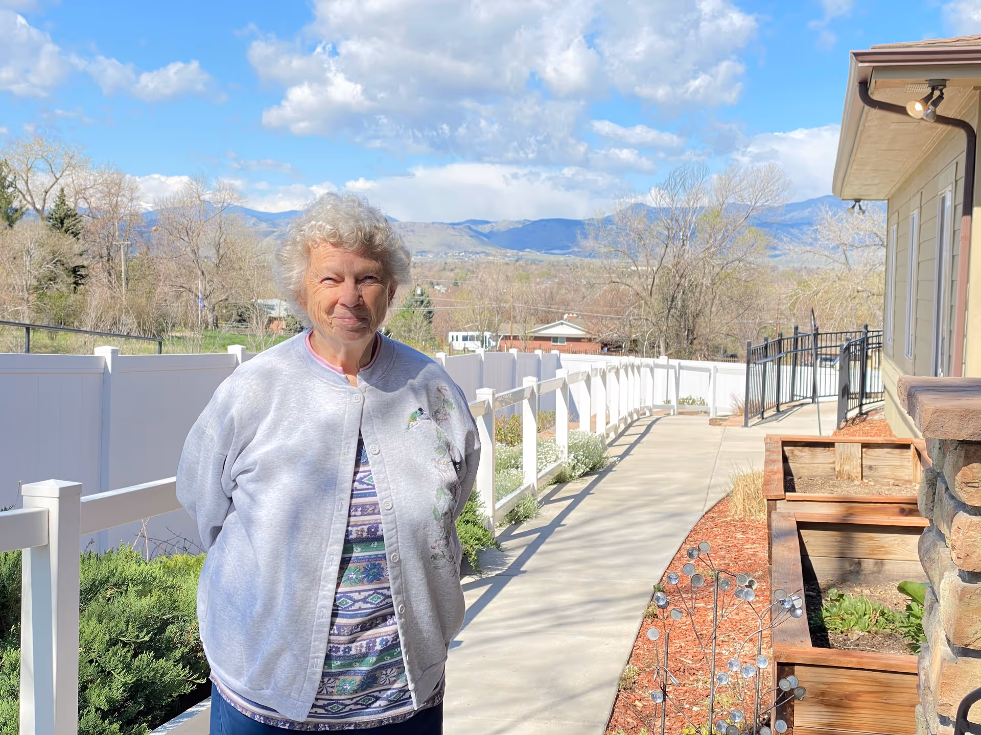 An older woman stands smiling on an outdoor paved walkway next to a building, with a white fence, landscaping, and distant mountains under a blue sky.