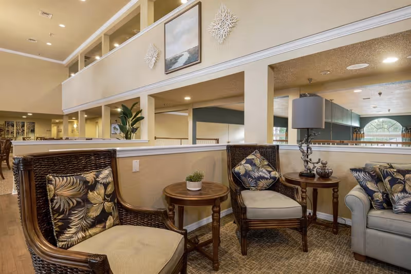 A cozy seating area in a senior living facility with two wicker armchairs featuring floral cushions, a small round wooden table with a potted plant, another wooden side table with a decorative lamp and ornaments, and a light gray sofa with matching floral cushions. The background shows a spacious, well-lit interior with beige walls, a large mirror, and artwork on the upper wall.