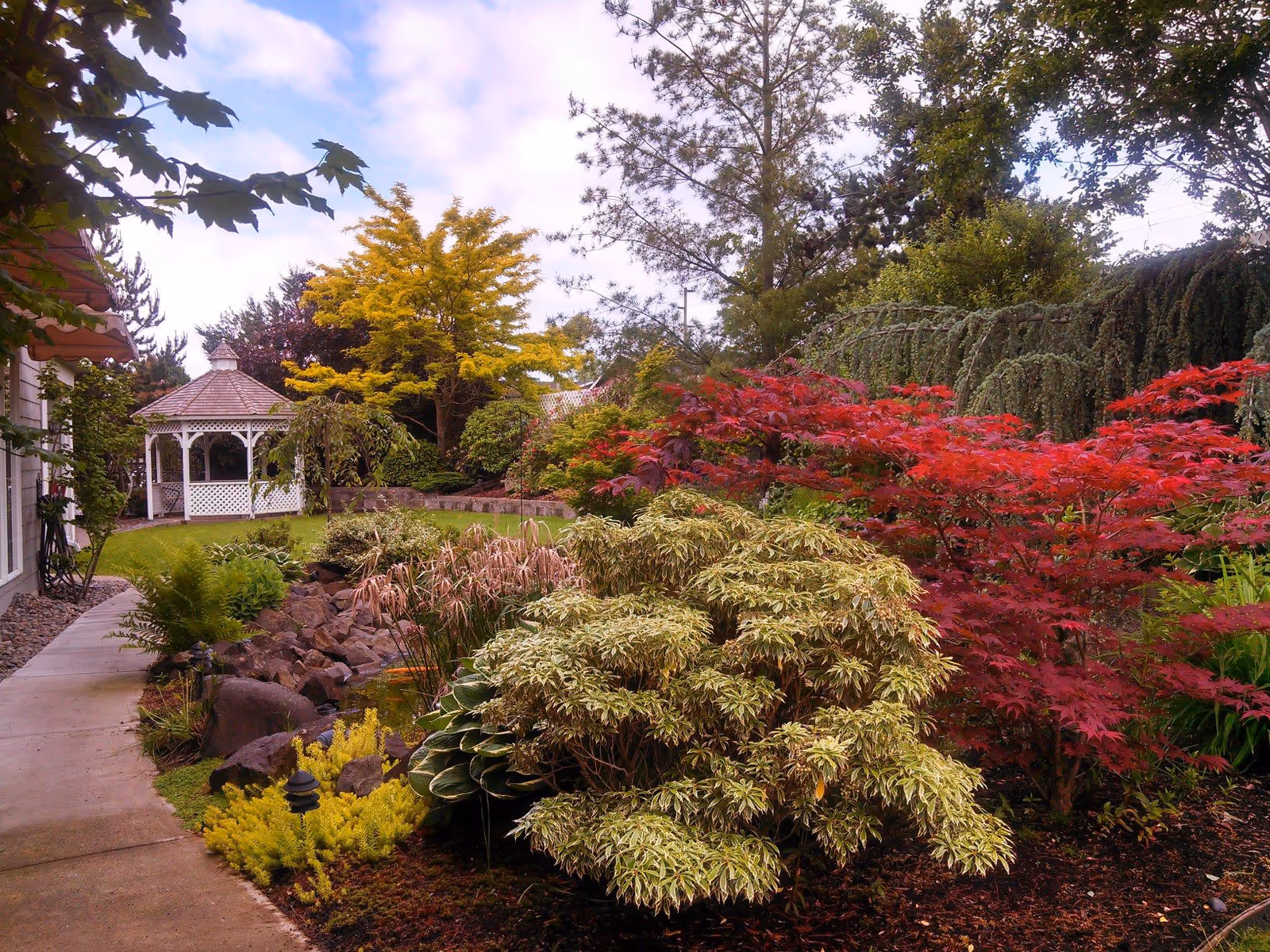 A landscaped garden area with a variety of colorful shrubs and trees, including red and yellow foliage. A white gazebo is visible in the background near a grassy lawn, and a concrete pathway runs along the left side of the garden.