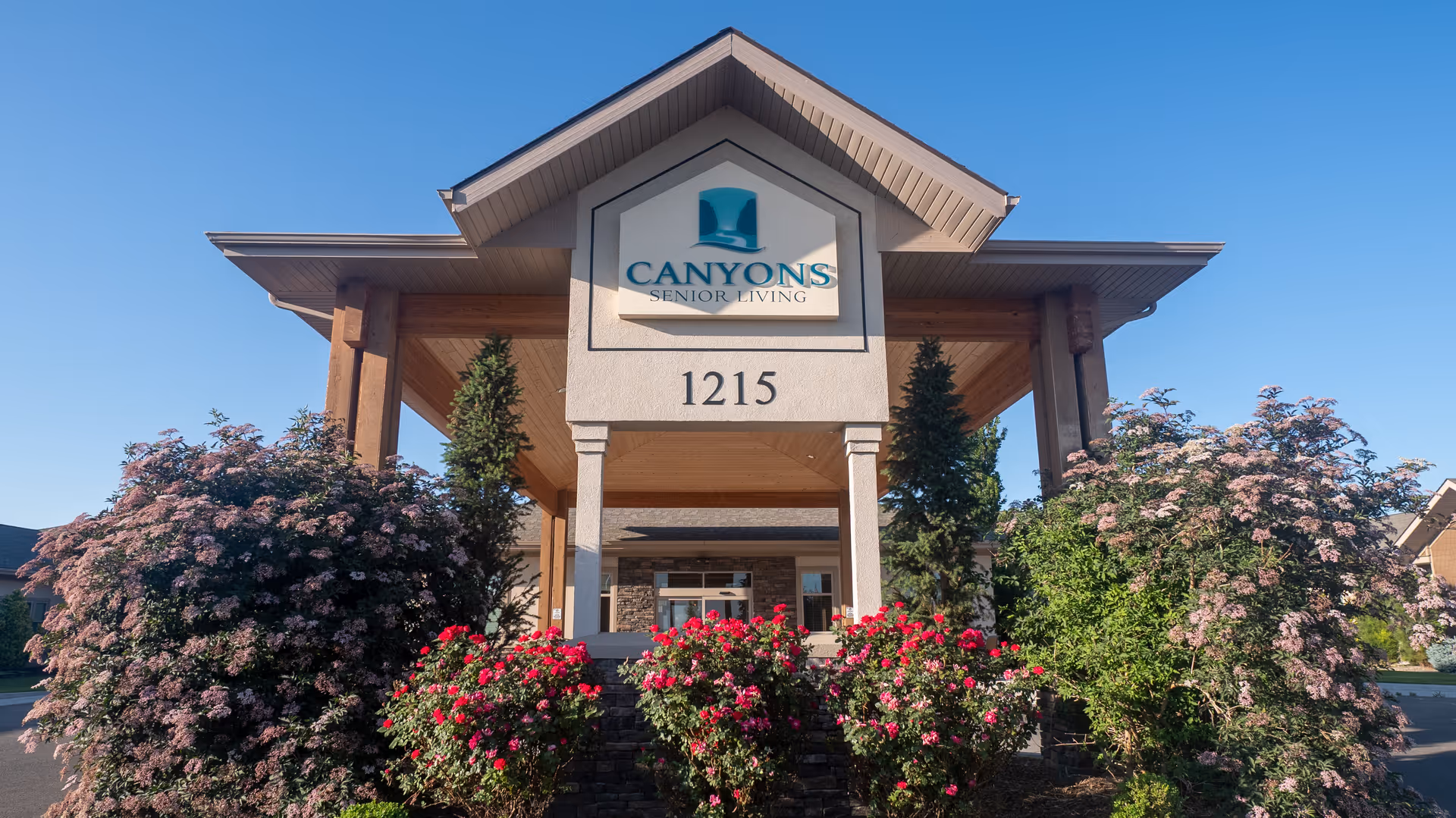 Front entrance of Canyons Senior Living facility with a covered porch supported by columns, surrounded by blooming bushes and trees under a clear blue sky.