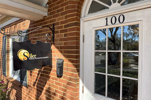 Exterior view of a brick building entrance with a white door numbered 100 and a hanging black sign that reads 'Siler Wealth Management'.