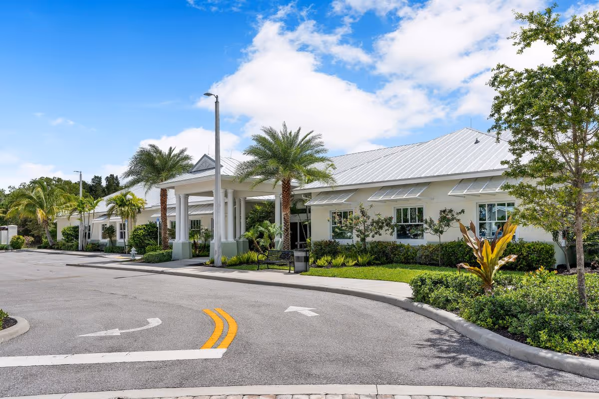 Exterior view of Luxe Senior Living at Jupiter facility showing a single-story building with white walls and a metal roof, surrounded by palm trees and landscaped greenery under a blue sky with some clouds. A curved driveway with directional arrows leads to the entrance with a covered portico supported by columns.