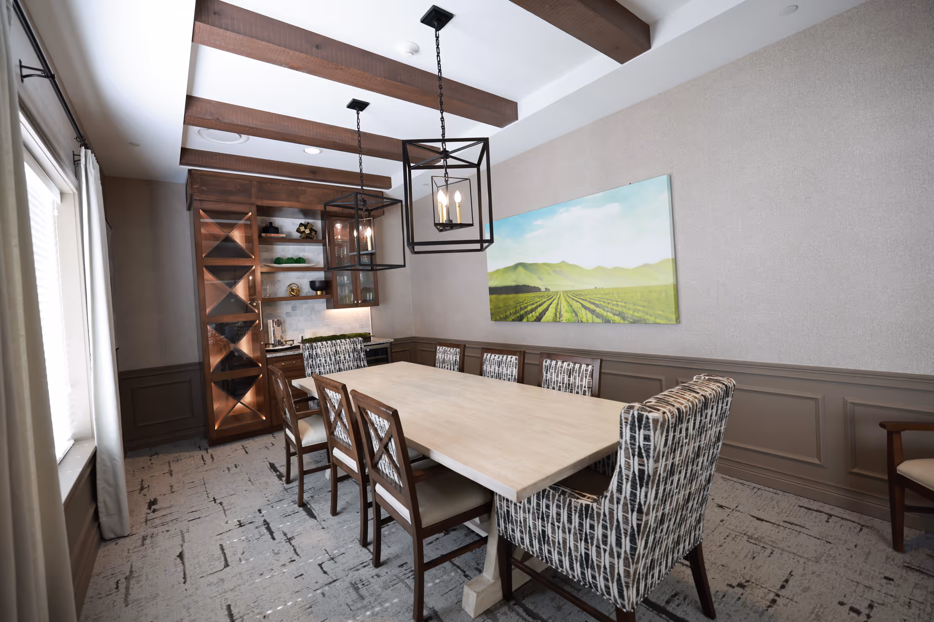 A dining room with a long rectangular wooden table surrounded by eight chairs, two of which have patterned upholstery. The room features wooden ceiling beams, two modern pendant lights hanging above the table, a large window with curtains on the left, and a cabinet with glass doors and shelves in the back corner. A landscape painting of green fields and mountains hangs on the right wall.