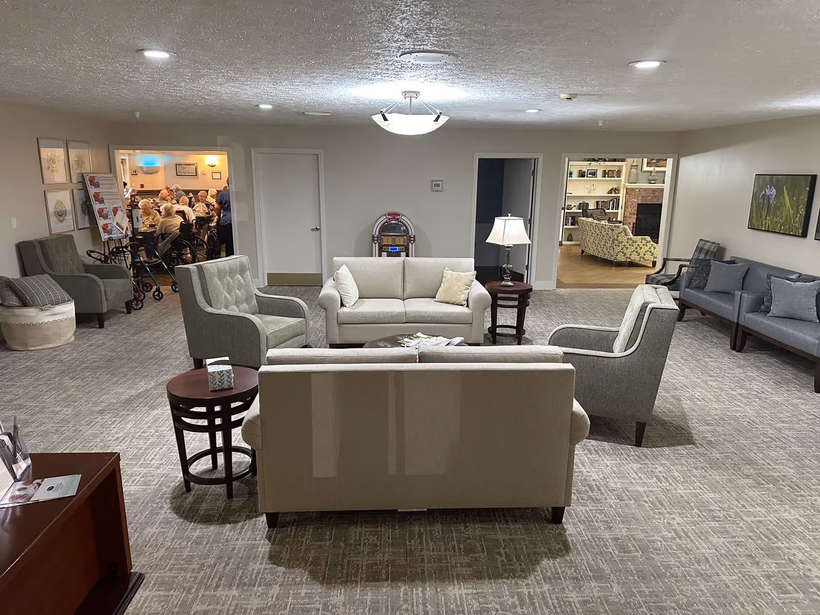 A spacious senior living facility common area with beige and gray sofas and armchairs arranged around a coffee table. There is a lamp on a side table and a jukebox against the far wall. The room has carpeted flooring and neutral-colored walls with framed artwork. Through open doorways, other rooms with additional seating and seniors gathered around tables are visible.