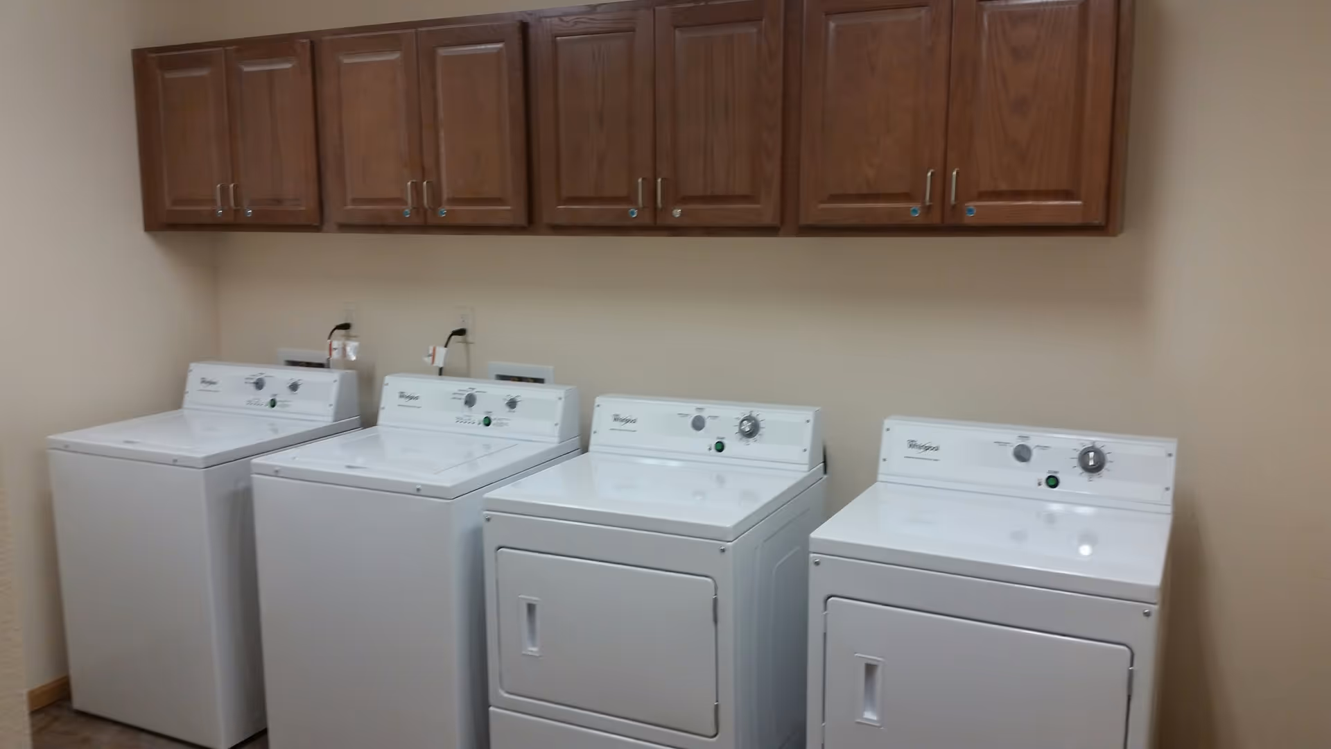 Laundry room with two washing machines and two dryers lined up side by side under a set of wooden cabinets mounted on a beige wall.