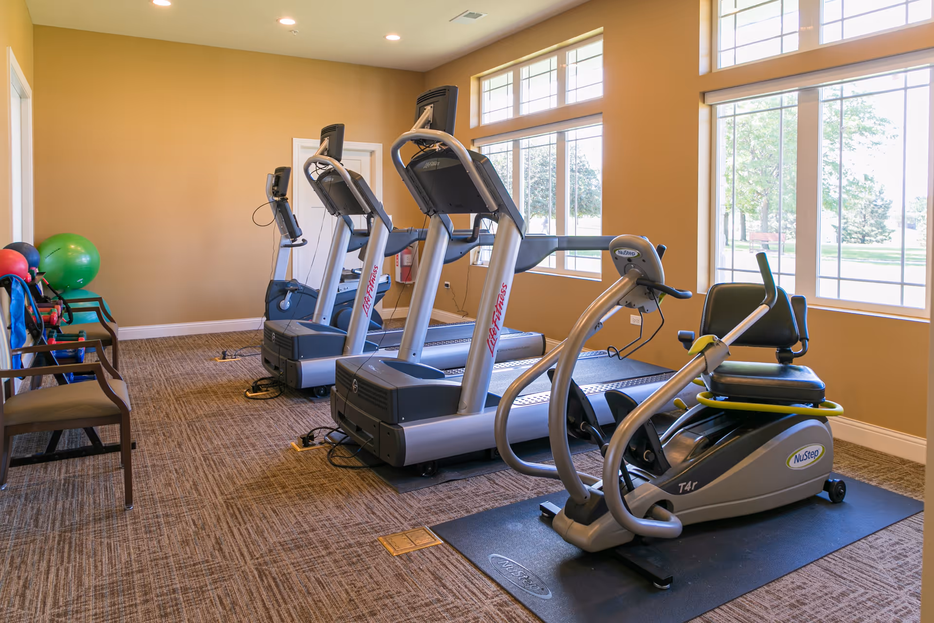 A fitness room with exercise equipment including two treadmills and a NuStep recumbent cross trainer. The room has large windows letting in natural light, beige walls, carpeted floor, and a few chairs along one wall with exercise balls nearby.