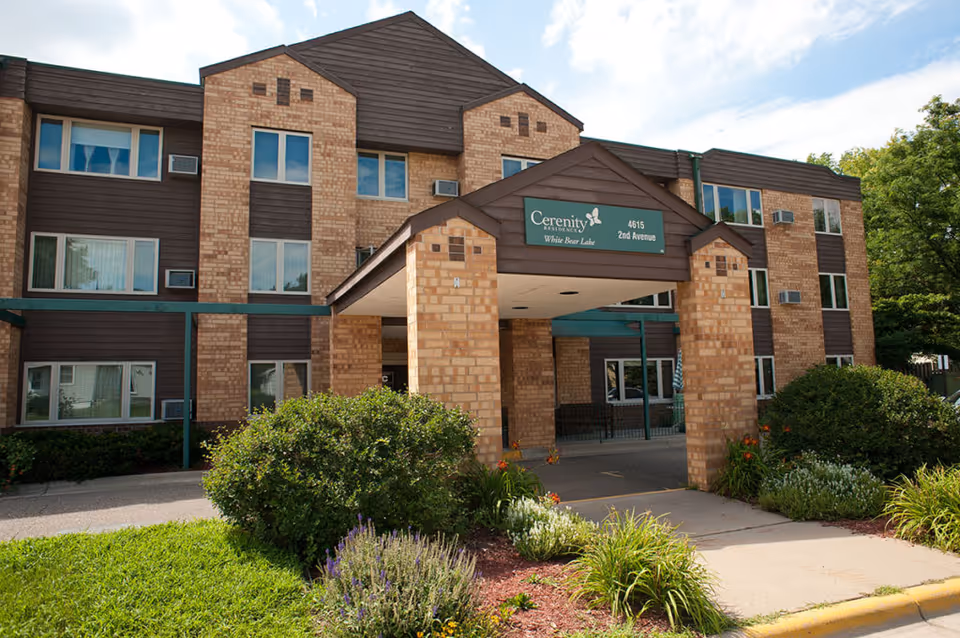 Exterior view of a three-story brick building with brown siding and multiple windows. The entrance features a covered portico with a sign that reads 'Cerenity Residence White Bear Lake 4615 2nd Avenue'. There are green bushes and landscaped plants in front of the building under a partly cloudy sky.