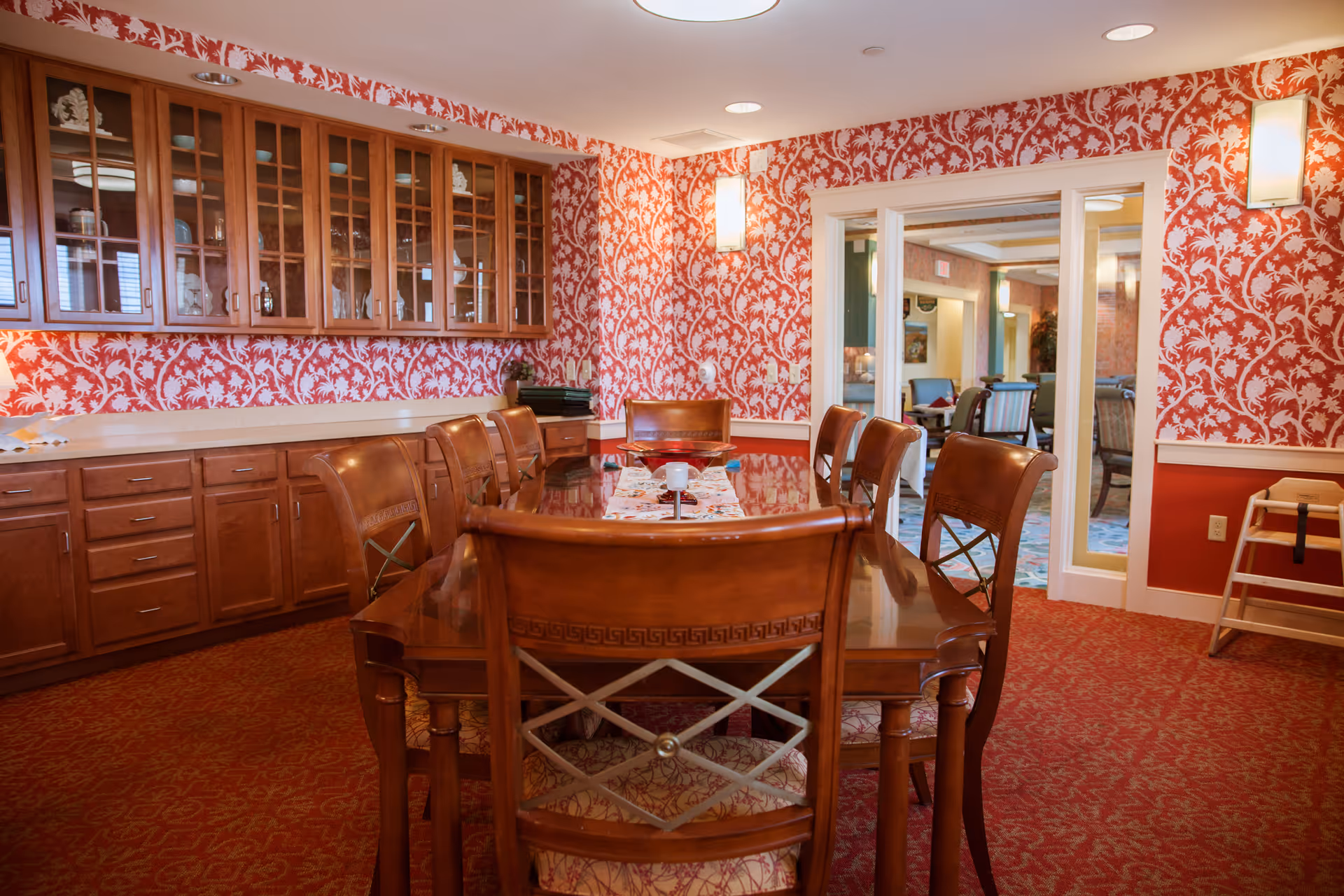 A dining room with a long wooden table surrounded by eight wooden chairs with cushioned seats. The walls are decorated with red and white floral wallpaper, and there are wooden cabinets with glass doors mounted on the wall. The room has red patterned carpet and two wall-mounted lights. A doorway with glass sliding doors leads to another room with additional seating.