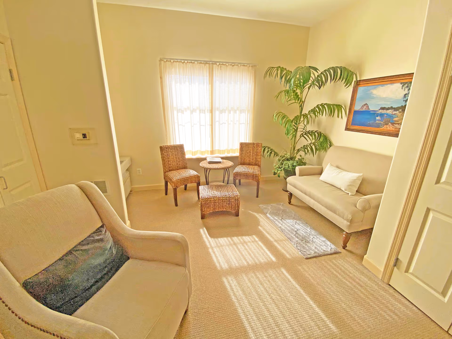 A bright living room with beige walls and carpet, featuring two beige upholstered chairs, a small round table with two wicker chairs, a wicker ottoman, a large potted plant, and a framed painting of a coastal scene on the wall. Sunlight streams through a window with sheer curtains.