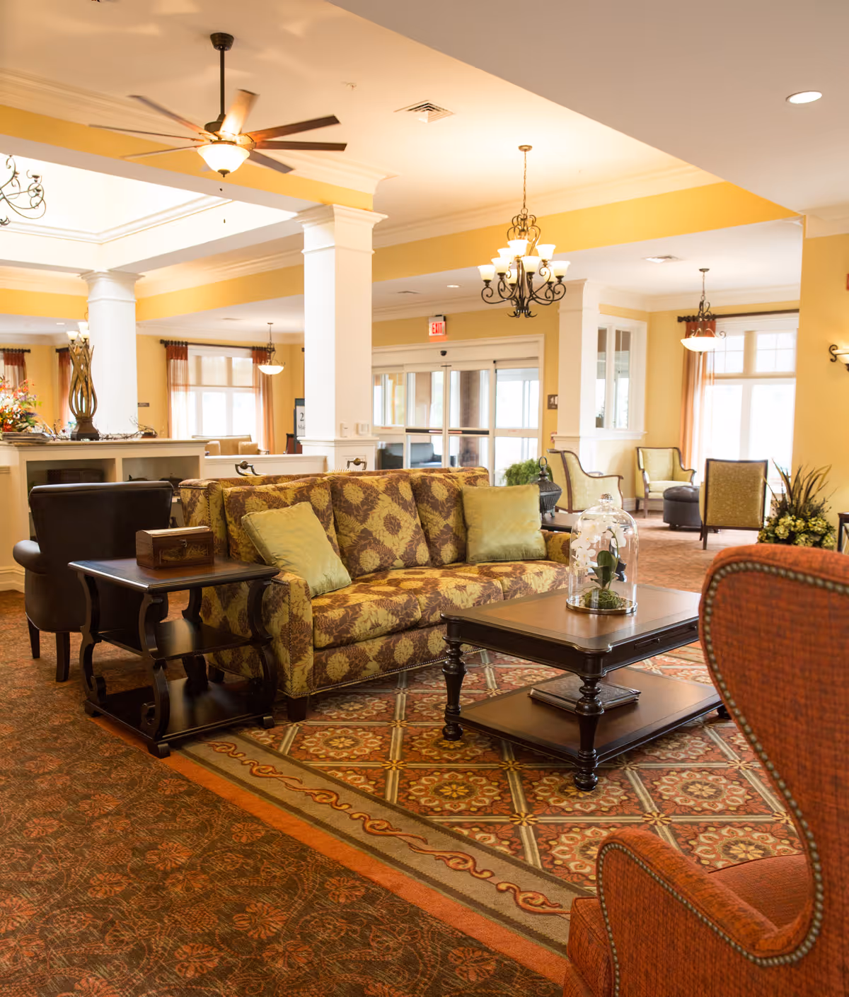 A spacious and warmly lit living room area in a senior living facility featuring a patterned sofa with green cushions, a wooden coffee table with a glass cloche containing a plant, an orange upholstered armchair, and additional seating in the background. The room has decorative chandeliers, ceiling fans, large windows with curtains, and a patterned carpet.