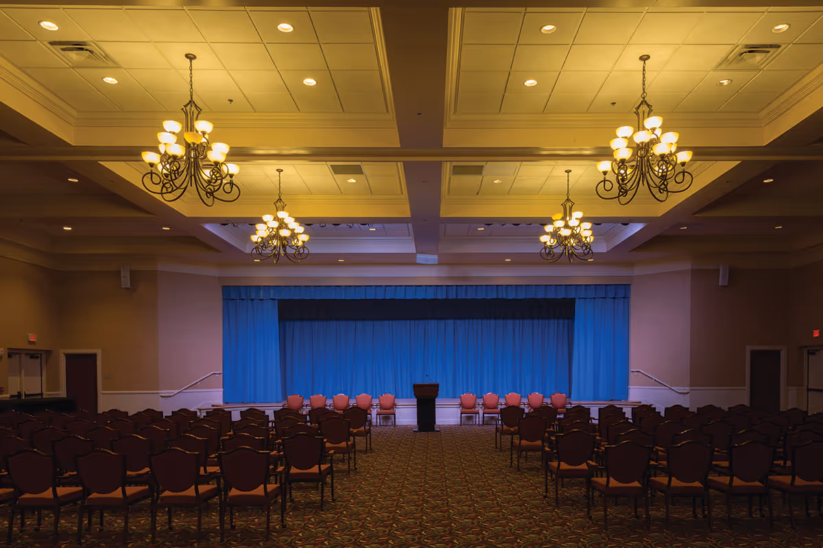 Large auditorium-style event room with rows of chairs facing a stage with blue curtains and chandeliers overhead.