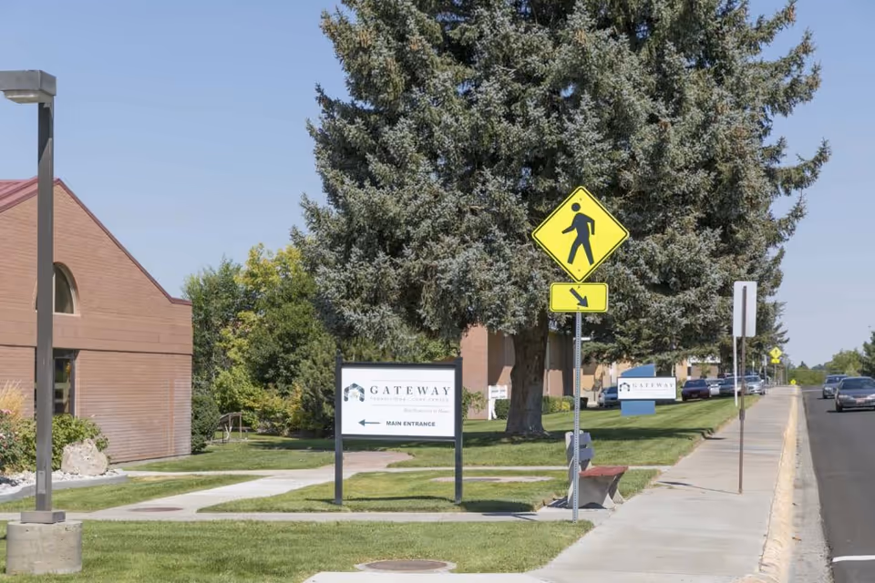 Exterior view of Gateway Transitional Care Center showing a sidewalk, a pedestrian crossing sign, a large tree, and the facility's main entrance sign on a grassy area next to a road with parked cars.