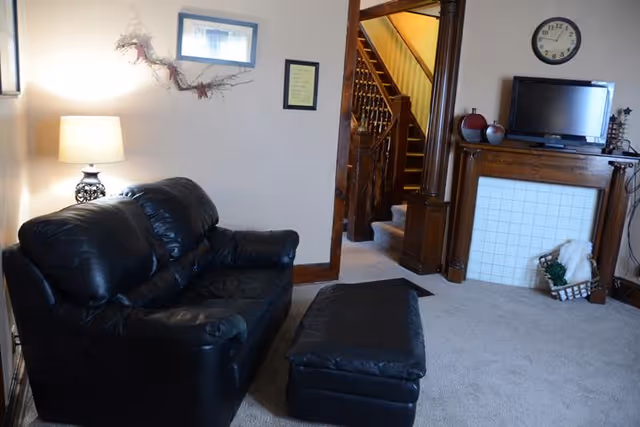A cozy living room with a black leather loveseat and matching ottoman on a carpeted floor. A table lamp with a white shade is on the left side, illuminating the space. On the wall above the loveseat are a framed picture and a decorative branch. To the right, there is a wooden fireplace mantel with a small TV on top, a clock on the wall above it, and decorative items including a basket with a blanket. In the background, a wooden staircase is visible through an open doorway.
