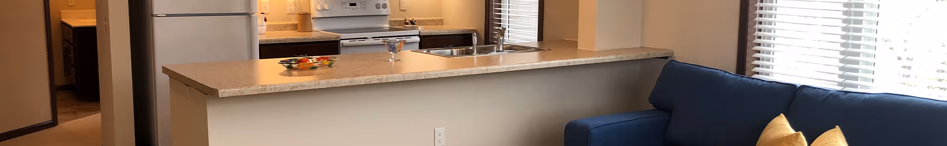 Open-plan interior showing a kitchen with an island countertop, stove and refrigerator alongside a blue sofa by a window.