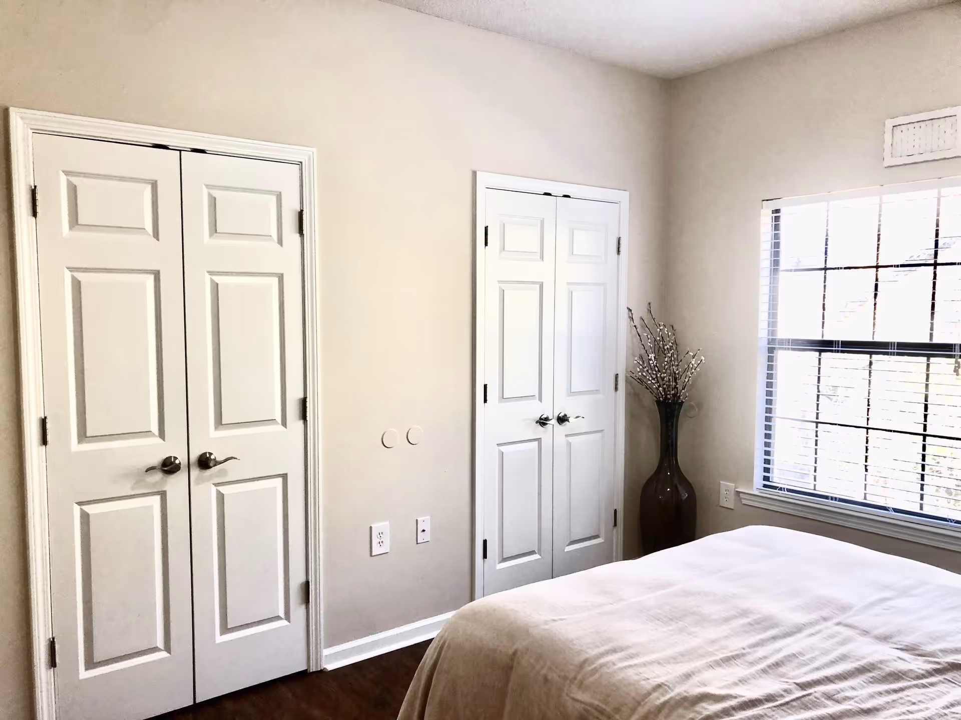 A bright bedroom with two white double-door closets, a large window with blinds, a bed with white bedding, and a tall vase with decorative branches in the corner.