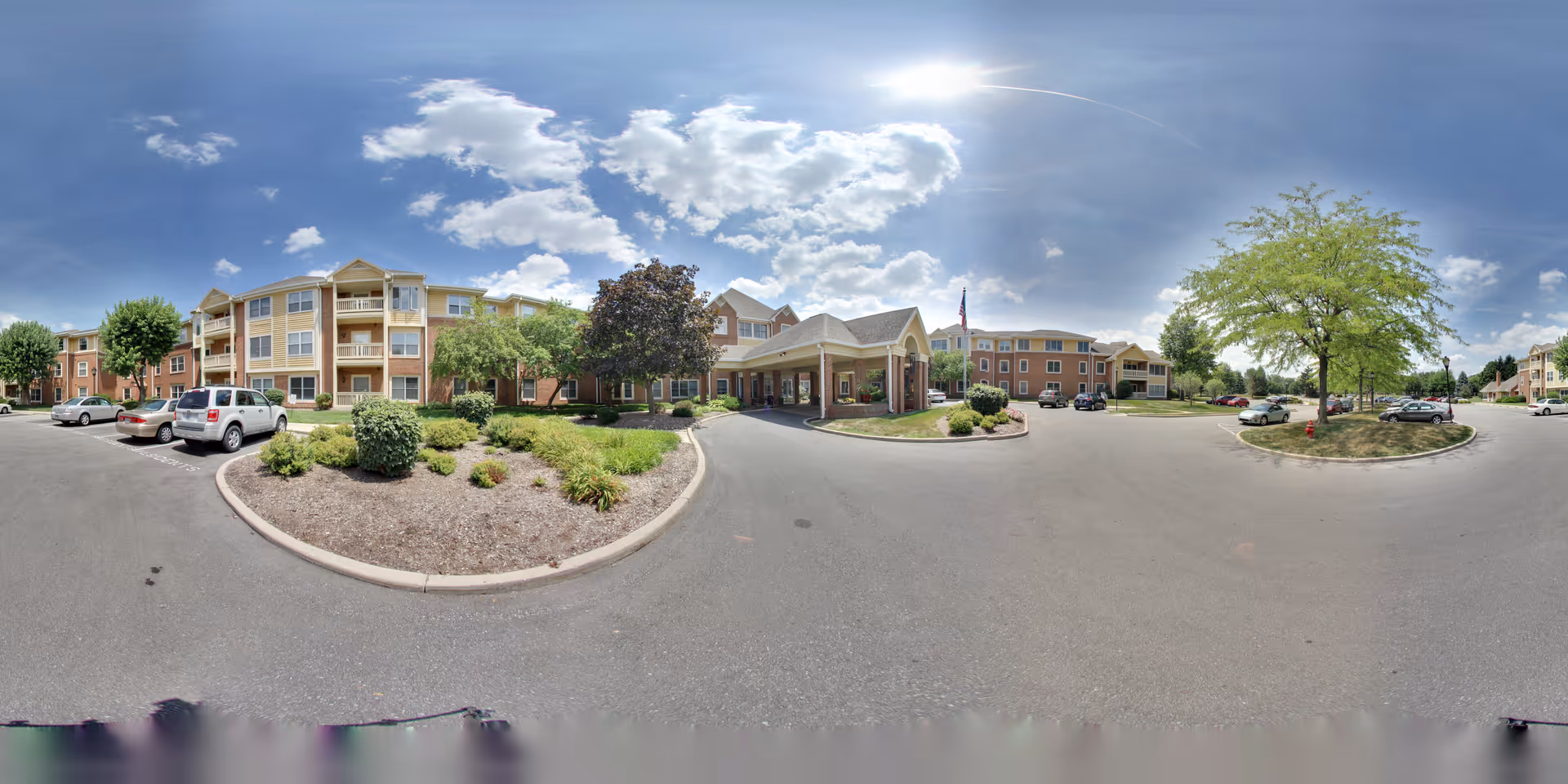 Wide panoramic view of the exterior of The Forum at the Crossing senior living facility showing a multi-story building with balconies, a covered entrance, landscaped garden beds, several parked cars, and a bright partly cloudy sky.