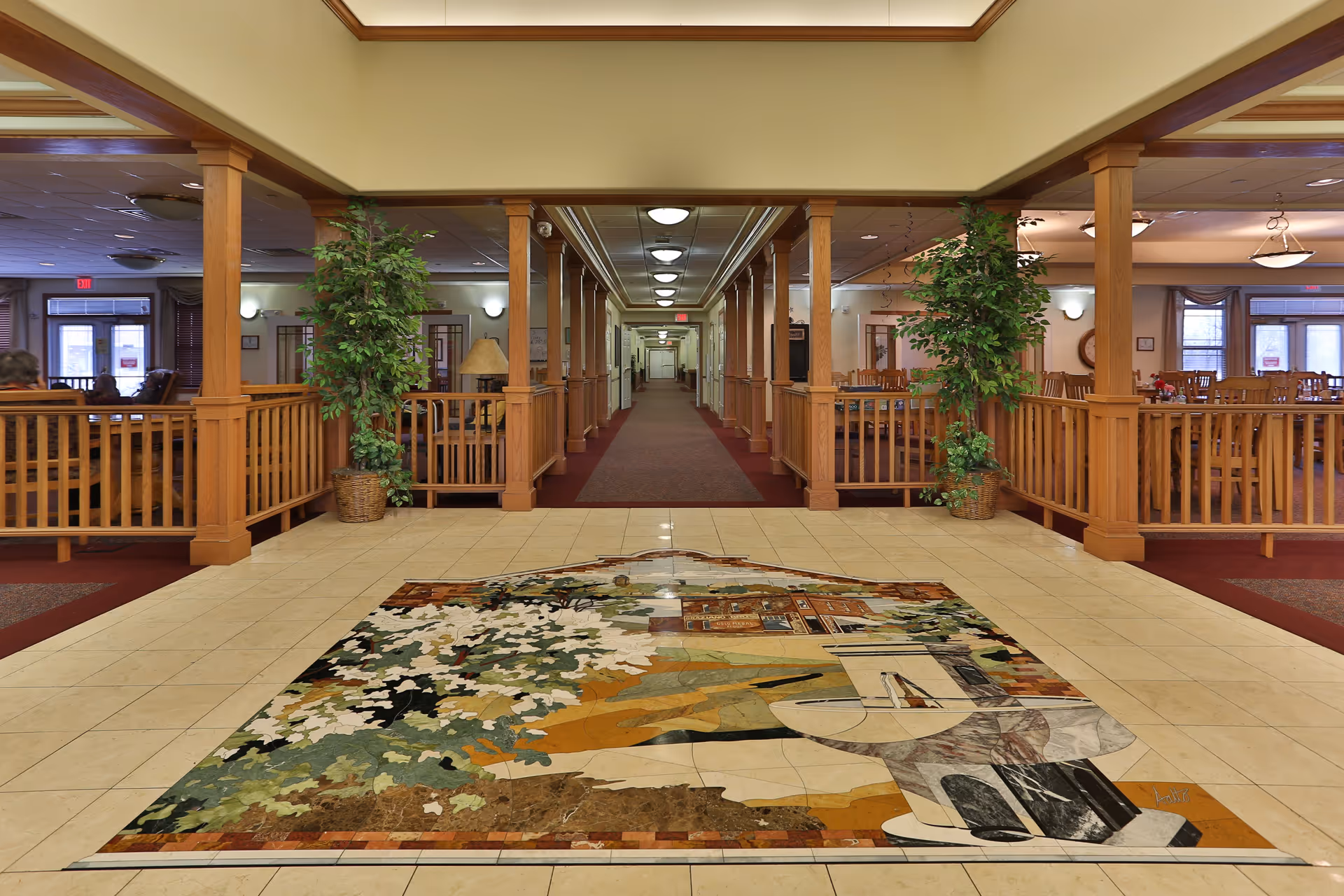 Interior view of a senior living facility hallway with wooden railings and columns on both sides. There are potted plants near the entrance, a decorative tile mosaic on the floor, and seating areas with tables and chairs visible on the right side. The hallway extends straight ahead with ceiling lights evenly spaced.