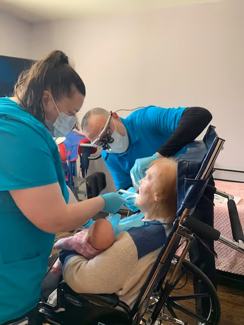 A healthcare professional and a dentist wearing masks and gloves are examining the mouth of an elderly woman seated in a wheelchair, who is holding a baby doll. The setting appears to be a clinical or care facility room with a bed in the background.