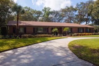 Single-story brick building with a brown roof surrounded by trees and greenery. A curved concrete driveway leads up to the building entrance under a partly cloudy sky.