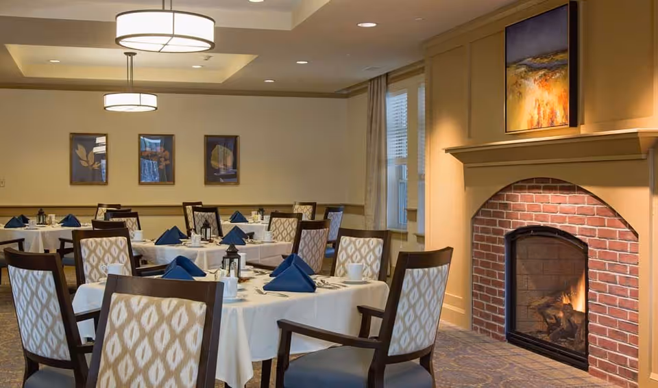 A dining room with several round tables covered with white tablecloths, each set with blue folded napkins, cups, and silverware. The room features patterned chairs, a lit brick fireplace with a colorful painting above it, three framed artworks on the wall, and two modern ceiling lights. There is a window with curtains on the right side.