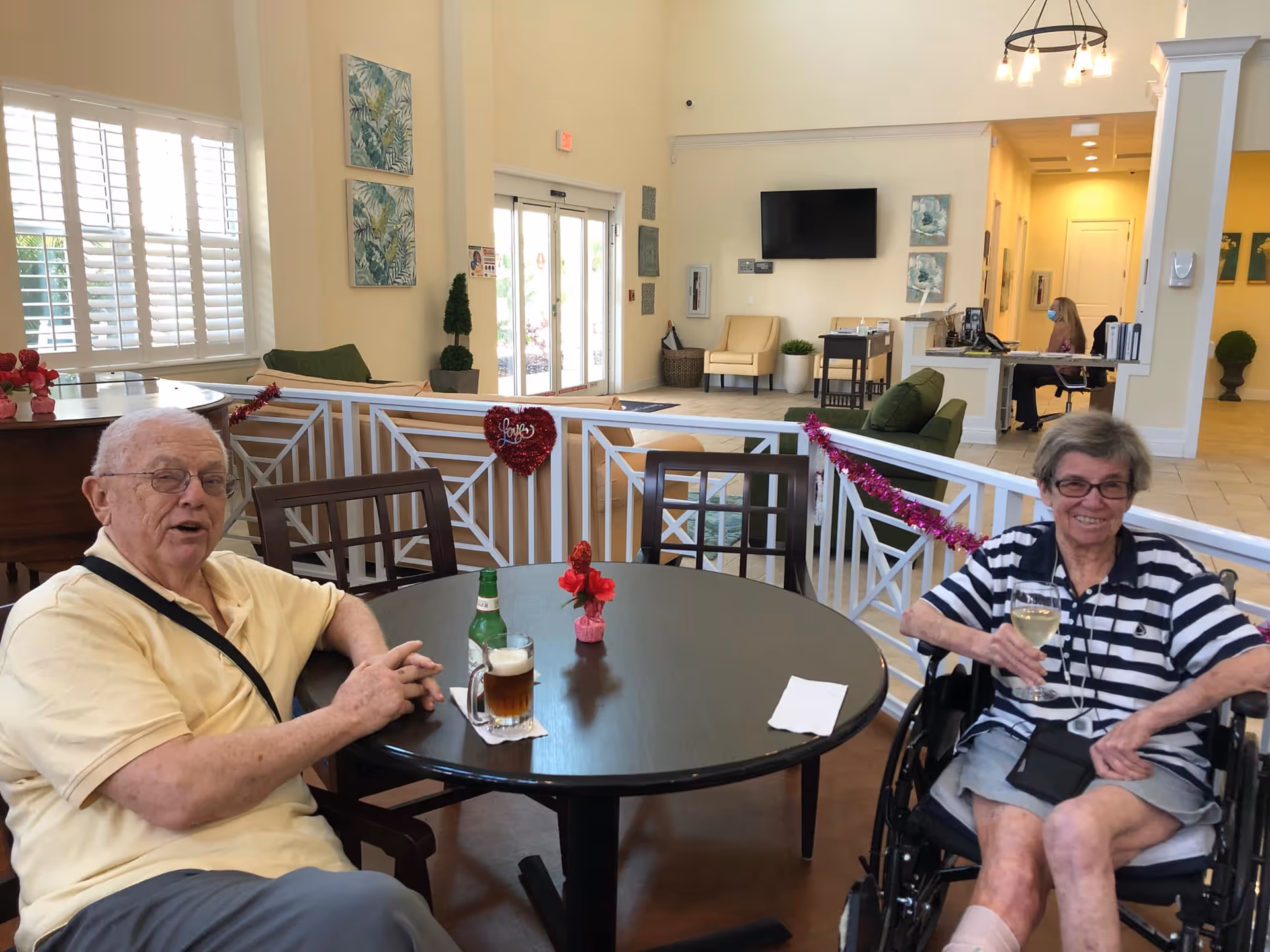 Two elderly individuals sitting at a round table in a bright, spacious common area of a senior living facility. One man is wearing a yellow shirt and glasses, holding a glass of beer, while the woman in a wheelchair is wearing a striped shirt and holding a glass of white wine. The background shows a reception desk, chairs, wall art, and large windows with white shutters.