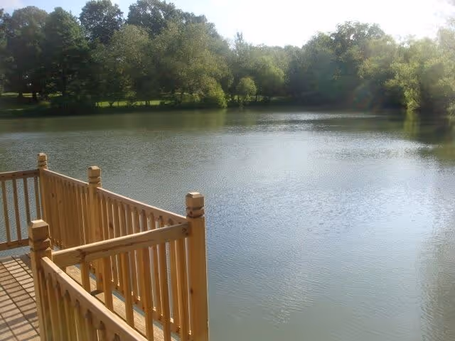 View of a calm lake surrounded by trees with a wooden railing in the foreground, suggesting a deck or pier overlooking the water.