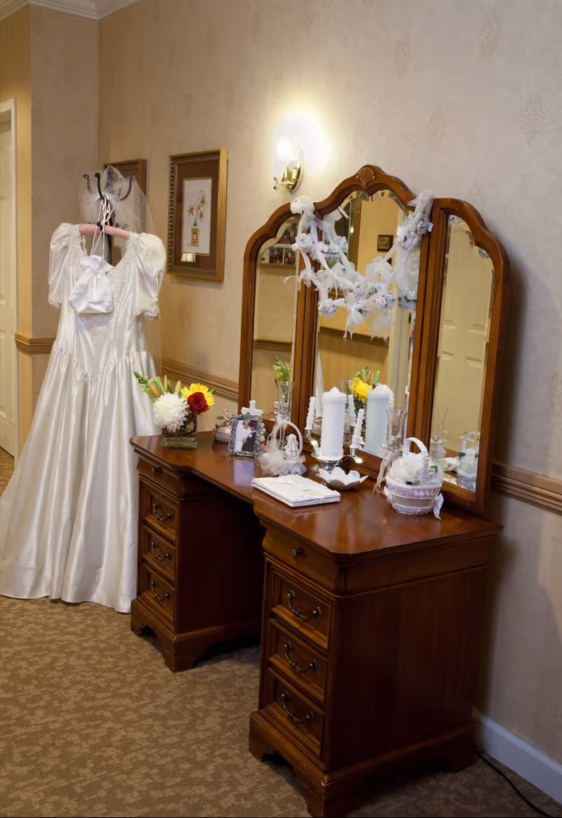 Wooden vanity with a three-panel mirror decorated with white garlands, a bridal gown on a hanger beside it, and candles, flowers, and framed photos on the tabletop.
