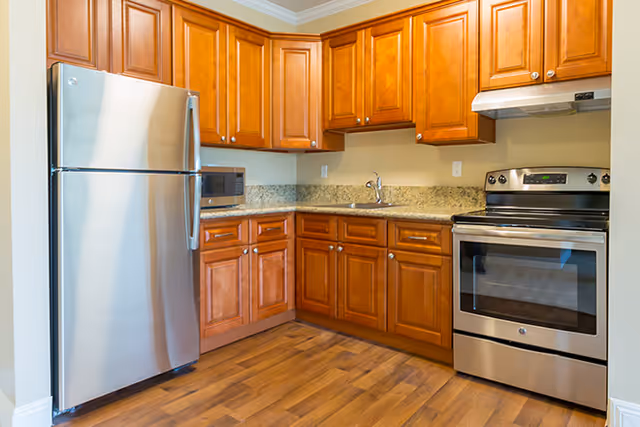 A kitchen with wooden cabinets, granite countertops, a stainless steel refrigerator, a microwave, a sink, and a stainless steel electric stove with an overhead vent hood. The floor is wood and the walls are light-colored.