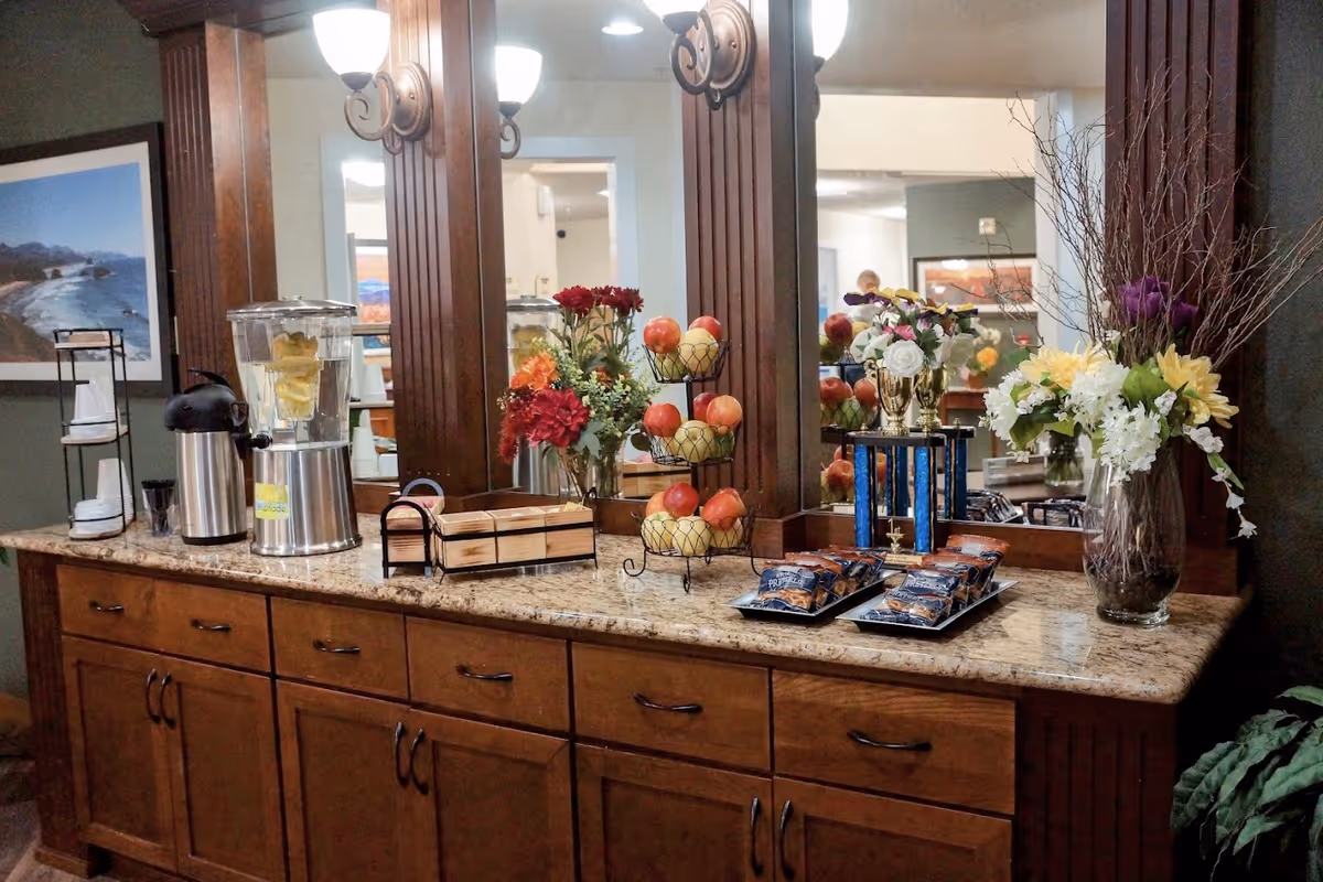A countertop with wooden cabinets below, featuring a water dispenser with lemon slices, a coffee dispenser, a tiered fruit basket with apples, a trophy, trays with packaged snacks, and two vases with colorful flower arrangements. The background includes large mirrors and wall sconces with lights.