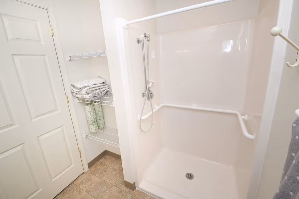 A white accessible shower stall with grab bars and a handheld showerhead next to shelves holding towels and toilet paper.