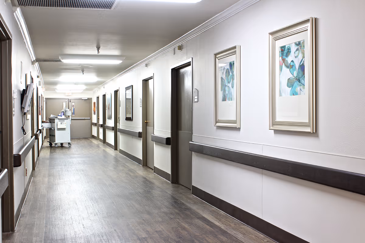 A clean, well-lit hallway in a healthcare facility with several closed doors on both sides, handrails along the walls, framed artwork, and a medical cart near the end of the corridor.