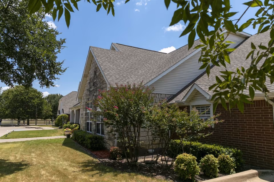 Exterior view of a senior living facility building with a combination of stone and brick walls, surrounded by green shrubs and trees under a clear blue sky.