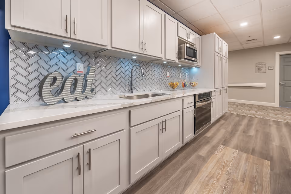 Long modern kitchenette with white cabinets, marble countertop, herringbone tile backsplash and stainless steel appliances.