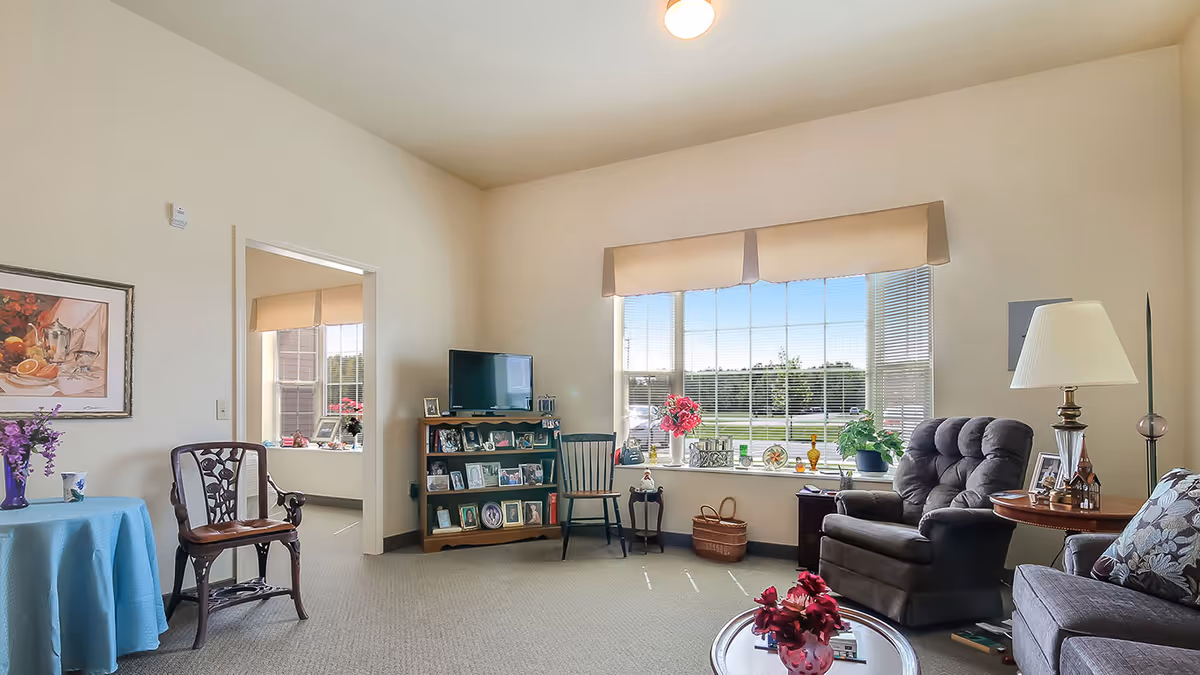 Bright assisted living facility living room with armchairs, a TV and photo shelf, coffee table with flowers, and a large window overlooking the grounds.
