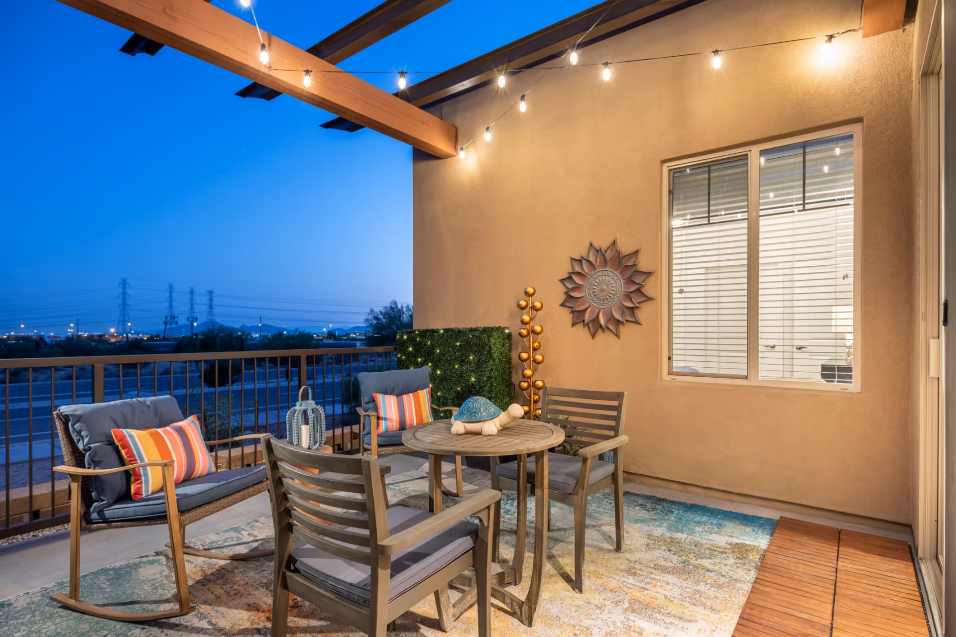 A cozy outdoor patio area at dusk with a round wooden table surrounded by four cushioned chairs, two of which have colorful striped pillows. The patio is decorated with string lights hanging from the wooden beams above, a decorative sun wall art, a small green hedge with lights, and a golden ornamental sculpture. The patio overlooks a distant view of power lines and a cityscape under a clear blue sky.