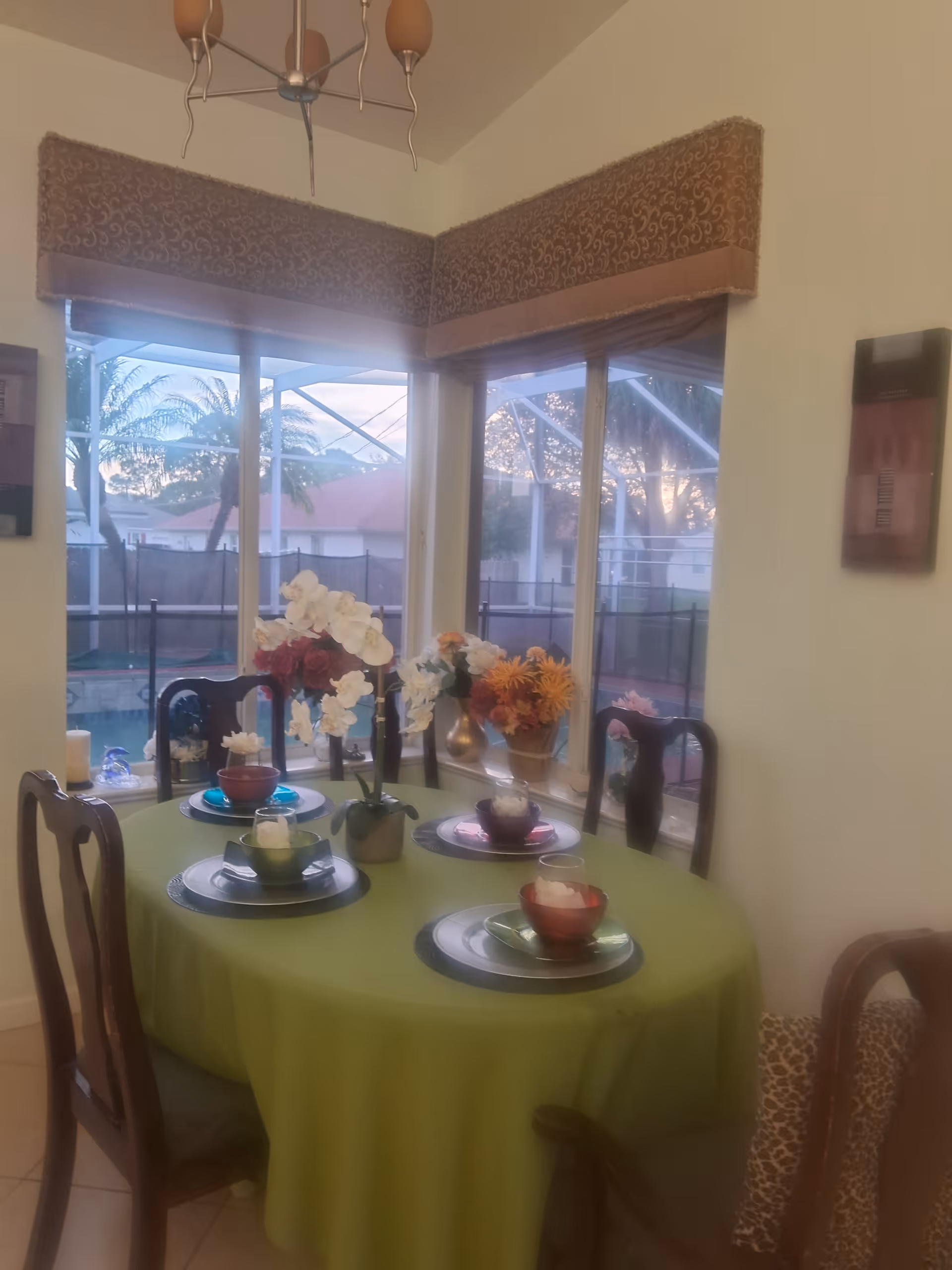 A dining room with a round table covered with a green tablecloth, set with plates, bowls, and glasses. There are five wooden chairs around the table. The room has large windows with a view of an outdoor pool area and palm trees. Floral arrangements are placed on the table and windowsill. A chandelier hangs from the ceiling.