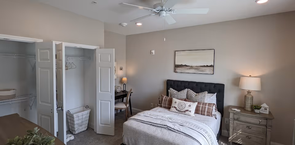 A cozy bedroom featuring a neatly made bed with multiple pillows, a bedside table with a lamp and small decorative items, an open closet with hangers and a laundry basket, a desk with a chair and lamp, and a ceiling fan above. The walls are painted light beige and there is a landscape painting hanging above the bed.