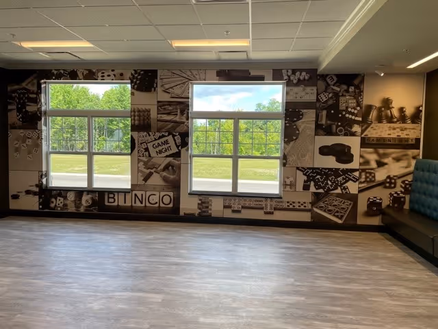 A spacious room with two large windows letting in natural light. The wall behind the windows is decorated with a black and white mural featuring various game-related images such as dice, dominoes, bingo cards, and game pieces. There is a cushioned bench along the right side of the room.