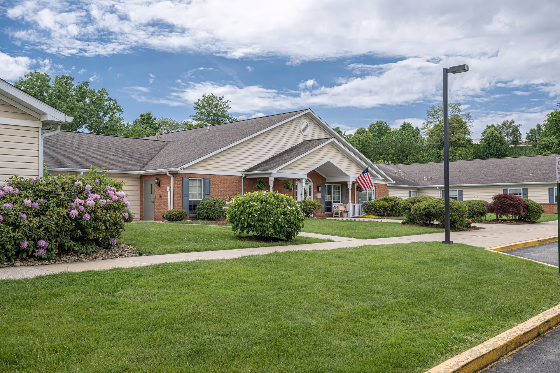 Exterior view of a single-story senior living facility building with beige siding and brick accents. The building has a covered entrance with white pillars and an American flag displayed. There are well-maintained green lawns, bushes, and flowering plants along the sidewalk leading to the entrance. A street lamp and a paved driveway are visible on the right side under a partly cloudy sky.