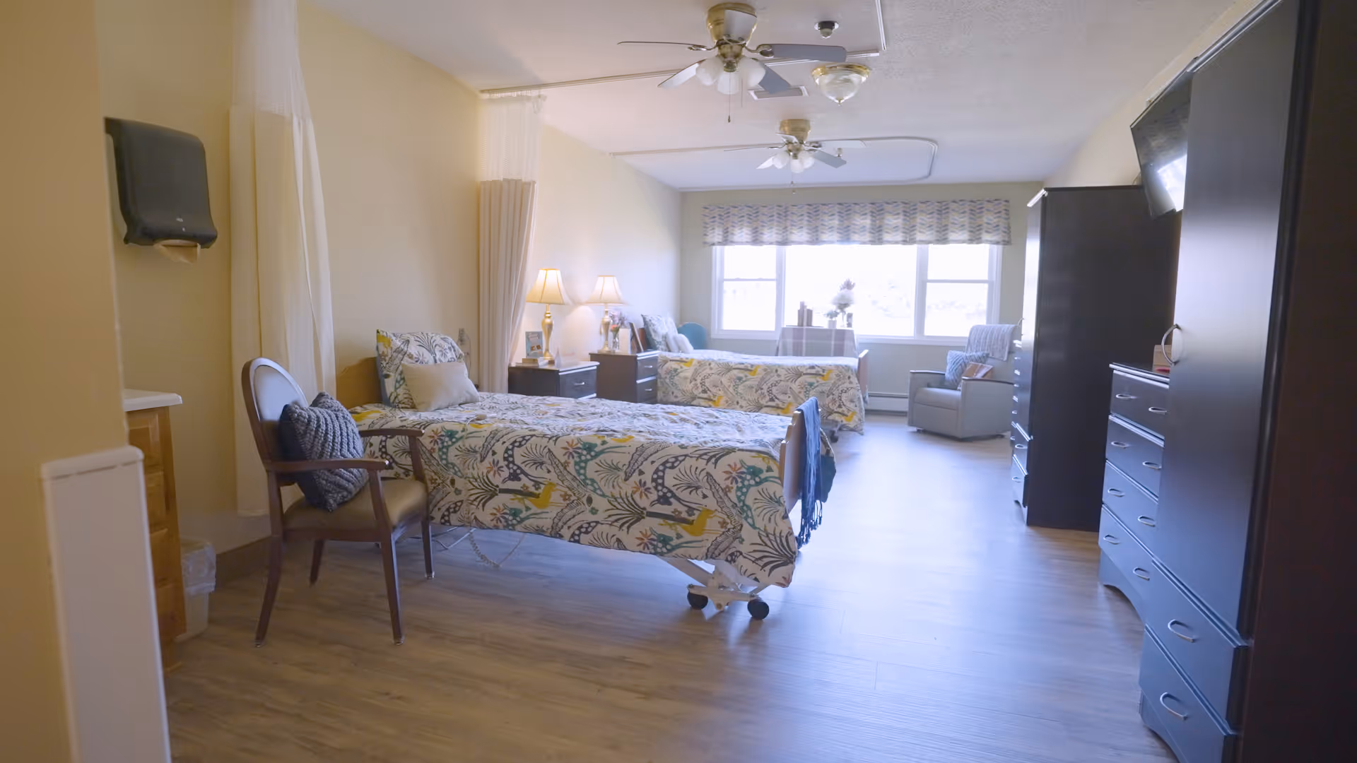 A bright nursing home bedroom with two hospital-style beds covered in floral patterned bedding. Each bed has a bedside table with lamps and personal items. There is a wooden chair with a cushion, a large window with patterned valance letting in natural light, and a recliner chair near the window. Dark wooden dressers and a TV mounted on the wall are also visible. The room has light-colored walls and wood flooring.