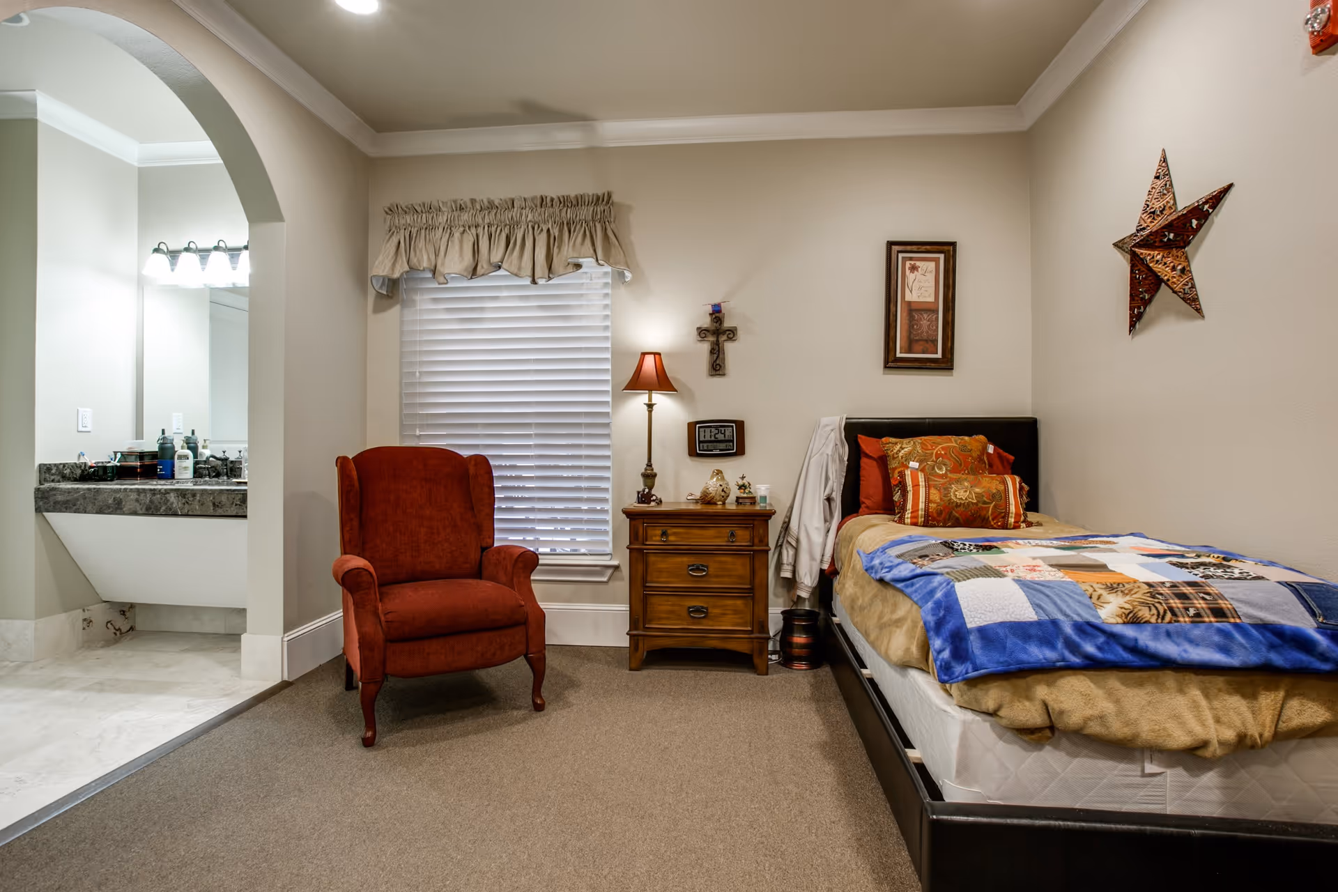 A cozy bedroom with a single bed covered in a colorful patchwork quilt and multiple pillows. Next to the bed is a wooden nightstand with a lamp, a clock, and decorative items. A red upholstered armchair is placed near a window with closed blinds and a beige valance. The room has beige walls adorned with a framed picture, a decorative cross, and a star-shaped wall hanging. An open archway leads to a bathroom area with a sink and mirror visible.