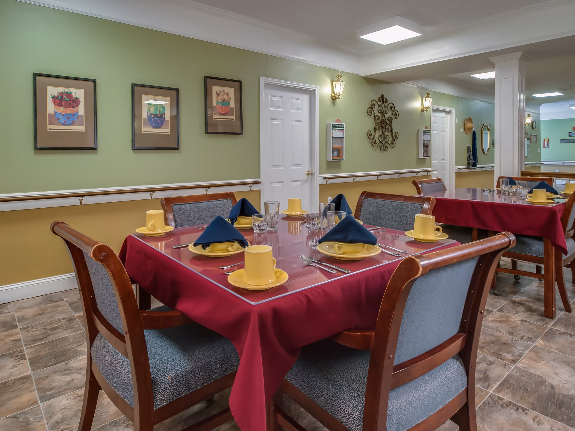 Dining area in a senior living facility with tables covered in burgundy tablecloths, set with yellow cups and saucers, blue folded napkins, glassware, and silverware. The room has green and yellow walls decorated with framed pictures and wall sconces, with wooden chairs upholstered in gray fabric around the tables.