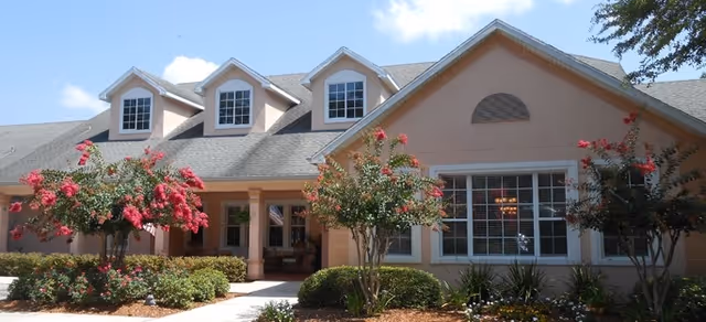 Exterior view of a single-story building with a light peach facade, multiple windows including three dormer windows on the roof, and a covered entrance. The front yard features neatly trimmed bushes, flowering trees with pink blossoms, and a clear blue sky above.