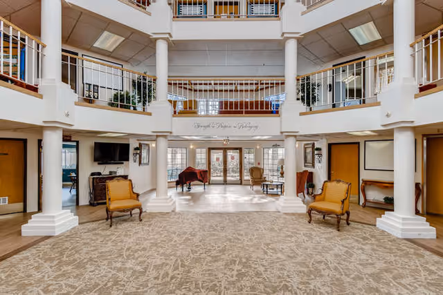 Spacious two-story lobby with columns, balcony railings, seating and a piano facing glass entrance doors.