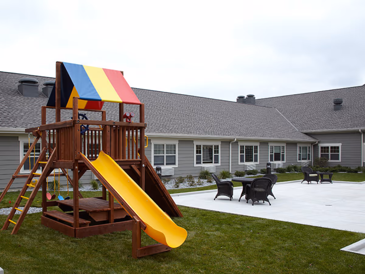 Outdoor play area with a wooden playground structure featuring a yellow slide, swings, and a colorful canopy. The playground is set on a grassy lawn next to a paved patio area with black wicker chairs and tables. The background shows a gray building with multiple windows and a shingled roof under a cloudy sky.