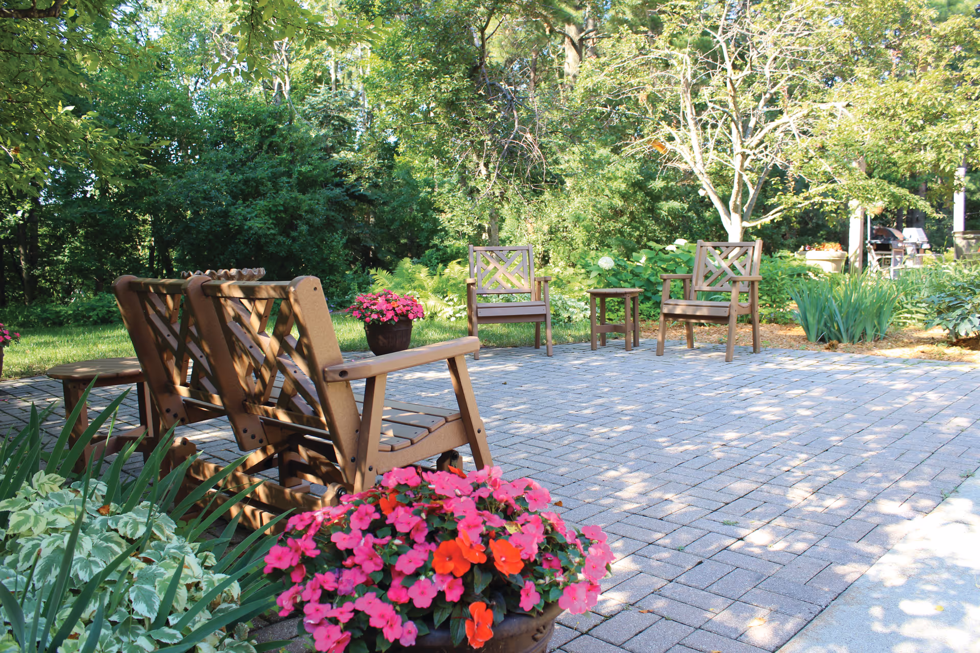 Outdoor patio area with several wooden chairs and small tables arranged on a paved surface, surrounded by lush green trees and plants. Bright pink and red flowers in pots add color to the scene, with sunlight filtering through the trees creating dappled shadows on the ground.