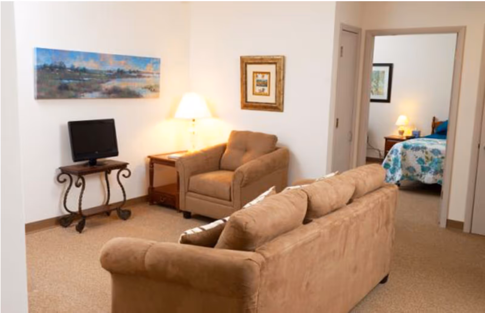 A cozy living room area with a brown sofa and armchair, a small TV on a decorative metal stand, a side table with a lamp, and framed artwork on the walls. Through an open doorway, a bedroom with a bed covered in a floral bedspread and a nightstand with a lamp is visible.