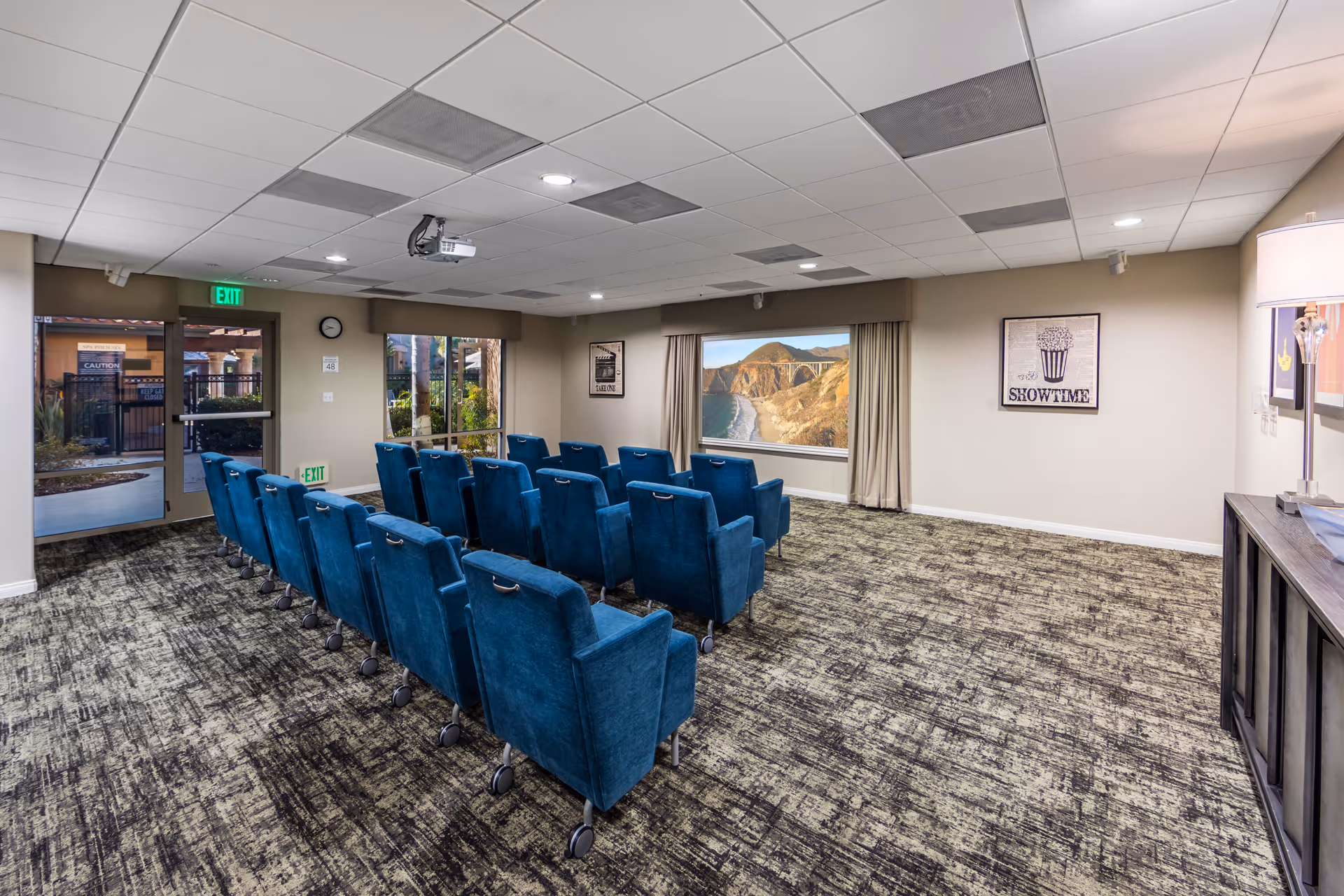A small theater room with three rows of blue upholstered chairs facing a large screen displaying a scenic coastal bridge and beach. The room has a patterned carpet, beige walls with framed artwork, a ceiling-mounted projector, and an exit door with windows showing an outdoor area.